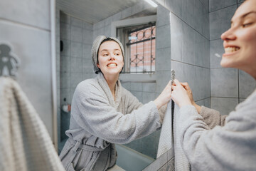 Woman in a cozy bathrobe brushing teeth and cleaning the bathroom mirror.