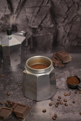 Geyser coffee maker with ground coffee standing on the table. Coffee in a black spoon. Pieces of broken dark chocolate. Spilled coffee beans. Dark background. Still life
