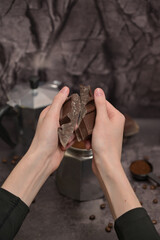 Woman's hands breaking pieces of dark chocolate. Geyser coffee maker with ground coffee standing on the table. Coffee in a black spoon. Spilled coffee beans. Dark background