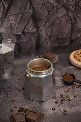 Geyser coffee maker with ground coffee standing on the table. Coffee in a black spoon. Pieces of broken dark chocolate. Spilled coffee beans. Oatmeal cookies on a wooden board. Dark background