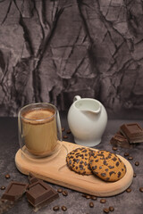 Coffee in a glass and oatmeal cookies on a wooden board. Milk jug on the table. Pieces of broken dark chocolate. Spilled coffee beans. Dark background. Coffee service