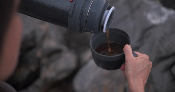 Over the shoulder close-up shot of hands pouring a hot drink from a thermos into a cup while sitting on rocks outdoors.