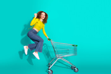 Young girl in casual yellow top and jeans posing playfully with a shopping cart against a vibrant...