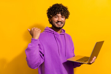 Young man with curly hair in a purple hoodie holding a laptop in front of a vibrant yellow...