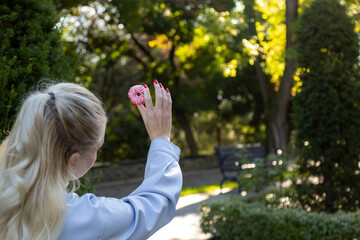 Rear view of blonde woman with red nails raising a pink frosted donut toward the golden sunlight while sitting in a peaceful, green park on a crisp autumn day