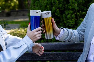Close-up of two people in light jackets clinking reusable thermos mugs one blue, one gold while sitting on a wooden bench in a green city park on a sunny autumn day