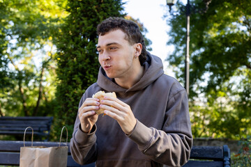 Young man in a brown hoodie sits on a wooden bench chewing a sandwich, smiling with full cheeks, surrounded by greenery and soft sunlight on a relaxed autumn day