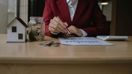 Fototapeta premium Businesswoman analyzing financial charts with a jar of coins and a small house model, representing investment, savings, and financial planning.