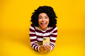 Cheerful young woman with curly hair and striped top excitedly holding a smartphone against a...
