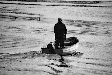 Black and white image of an experienced seafafer standing in his small boat, steering it out to sea in a timeless maritime scene.