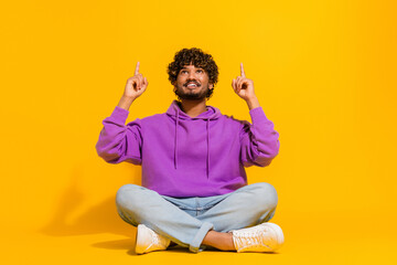 Young man in a purple hoodie sitting on a yellow background pointing upwards, representing casual...