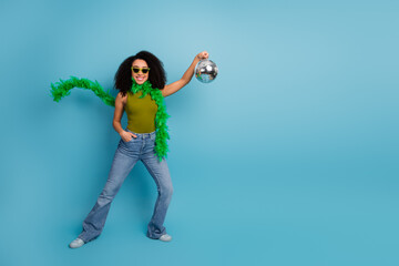 Joyful woman wearing green top and glasses holding a disco ball while posing against a vibrant blue...