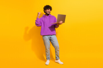 Funny man wearing a purple hoodie holding a laptop celebrates success on a vibrant yellow...
