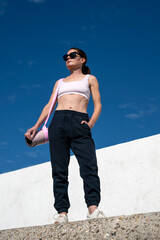 sporty woman in sports bar and jogging bottoms and exercise mat standing by a white wall in the...
