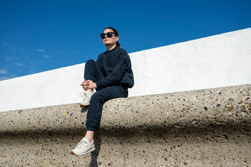 stylish woman sitting on a low concrete ledge, wearing a  tracksuit and sunglasses. Bright blue sky...