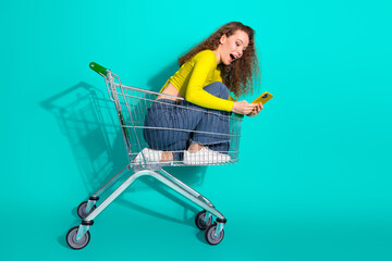 Young woman sitting playfully in a shopping cart on a teal background holding a mobile phone...