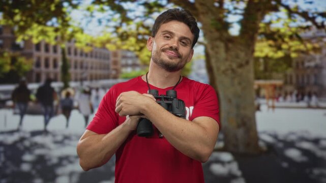 Man in red shirt holds binoculars to chest on street beside a plane tree and blurred crowd, smiling and relaxed; watchfulness duty serenity.