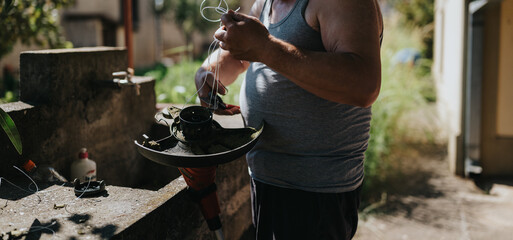 A man wearing a gray tank top works on a garden trimmer, handling wires and components outdoors. The scene conveys DIY home repair, outdoor work, and hands-on maintenance for garden tools.