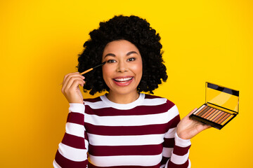 Happy woman applying makeup with a brush while holding an eyeshadow palette on yellow background
