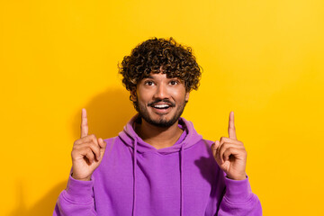 Smiling young man in purple hoodie against yellow wall making pointing signs with fingers showing...