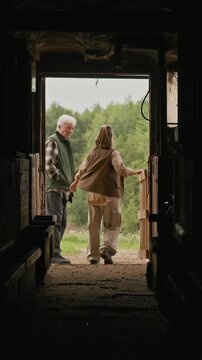 Vertical shot of young Caucasian female rancher closing wooden gate of farm shed, talking to senior man standing nearby and walking away together