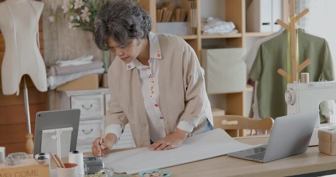 female tailor carefully drafting clothing pattern by hand on craft paper using sewing template at work table in sewing studio, 4k video.