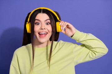 Excited young woman in yellow headphones enjoying music on purple background wearing casual...