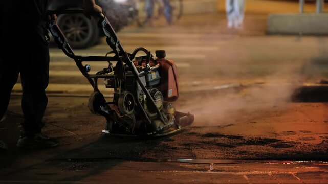 Worker compacting asphalt patching road at night during urban street repair