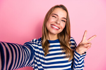 Portrait of smiling young woman making peace sign gesture against pink backdrop wearing striped...