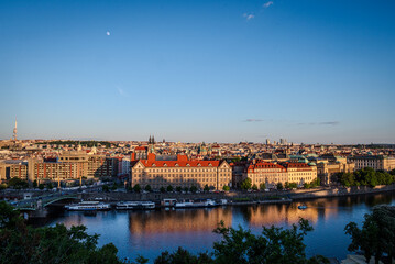 Evening sunlight bathes Prague Old Town, Czech Republic, with the Vltava River below and the moon rising in a clear sky- Horizontal