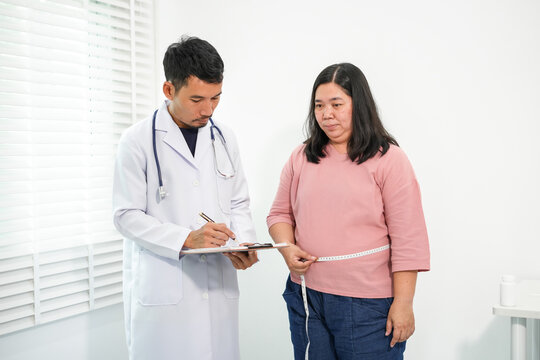 A doctor in a white gown and stethoscope records data on a clipboard while an Asian woman uses a tape measure to measure her waist for a medical consultation focused on health and weight.