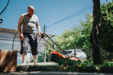 A senior man in a gray tank top and dark shorts mows a sunny backyard lawn with an electric lawn mower. Relaxed outdoor activity amid trees, fence, and clear blue sky.