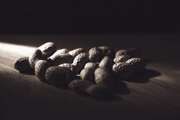 Pile of unpeeled peanuts illuminated by a single spot of light on a dark wooden table. Moody and dramatic food photography, black and white effect