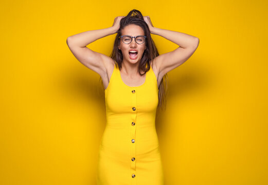Upset young woman with hands on head and tearing hair out, isolated on yellow background. Frustrated girl screaming.