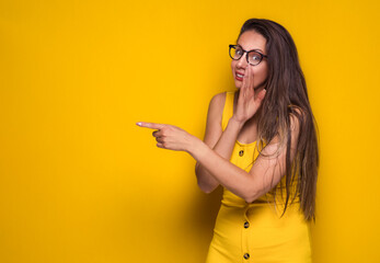 Psst. Young girl holding hand near mouth telling secret while pointing to something, isolated on yellow background. Copy space. Gossip concept.