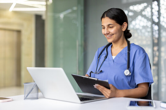 Young happy female doctor wearing scrubs and a stethoscope, smiling and writing on a clipboard while looking at a laptop, providing online medical care and virtual healthcare service