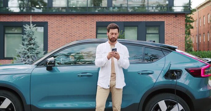 Serious young man in casual clothes using smartphone while standing next to electric car charging at urban station. Sustainable transport concept. Modern city lifestyle