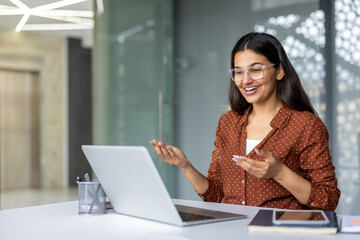Young smiling woman using laptop for online video call, communicating with colleagues during virtual meeting in modern office setting, actively engaging in discussion