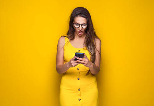 Impatient young woman with the mobile phone in the trouble, no time concept, isolated on yellow background. Worried girl looking to phone.