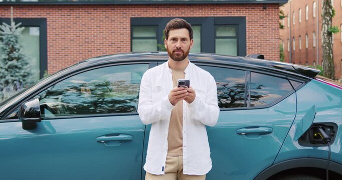Adult businessman in casual suit smiling and looking at camera while using smartphone next to his electric car charging at a city station, highlighting eco-friendly lifestyle and modern technology