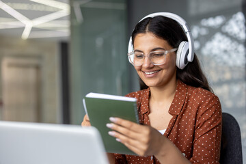 Young woman with glasses and headphones smiles while taking notes in a green notebook and engaging with an online course, on her laptop during remote learning at home