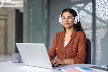 Young professional woman with white headphones typing on a laptop at a bright modern desk, focused on remote work and online learning in a contemporary office setting