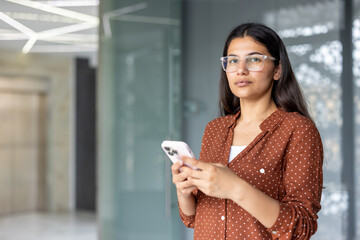 Indian woman wearing clear frame glasses and a polka dot shirt, holding a white smartphone, looking at the camera, conveying business connectivity and technology use in a modern office environment