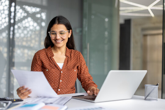 Young indian businesswoman sitting at her office desk, smiling while reviewing paper documents and typing on a laptop, representing efficiency and modern professional life - Powered by Adobe