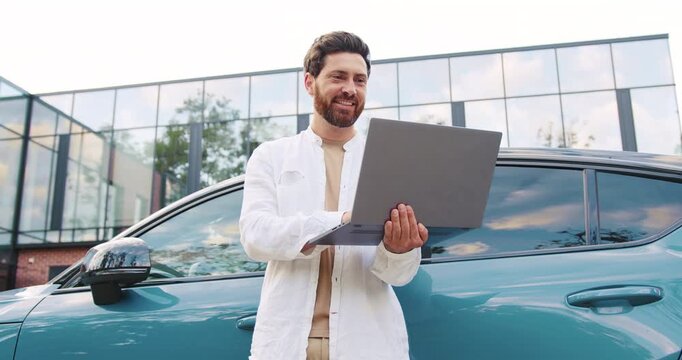 Caucasian man working remotely on laptop while standing next to his parked car outside modern office building, appearing focused and engaged in his task, exemplifying the flexibility of mobile work