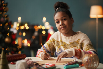 Portrait of Black girl teenager sitting at table writing holiday card with pencil, surrounded by...