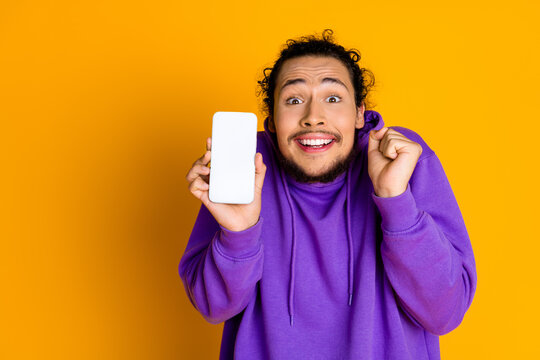Excited young man in purple hoodie happily shows smartphone screen against yellow background