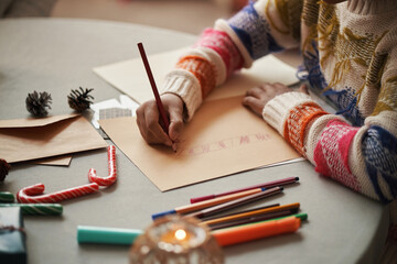 Caucasian child drawing Christmas card using colored pencils at table, hands visible creating festive artwork surrounded by candy canes, pine cones, envelopes, holiday decorations