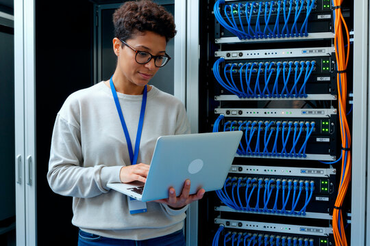 Young adult Black woman working on laptop while standing in server room, managing network cables and equipment, wearing glasses and identification badge, focusing on IT infrastructure