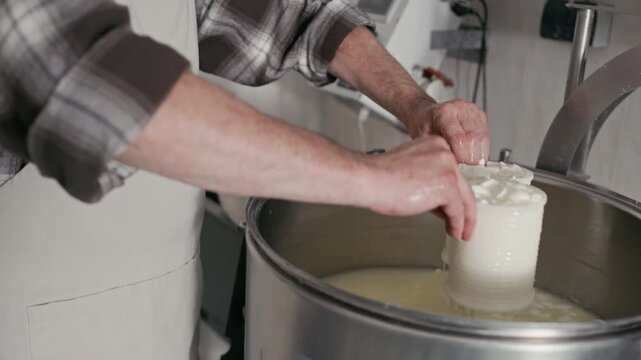 Cropped of hands of unrecognizable cheese factory worker in apron filling plastic molds with freshly cooked curd in metal vat and draining off whey carefully, crafting artisan cheese